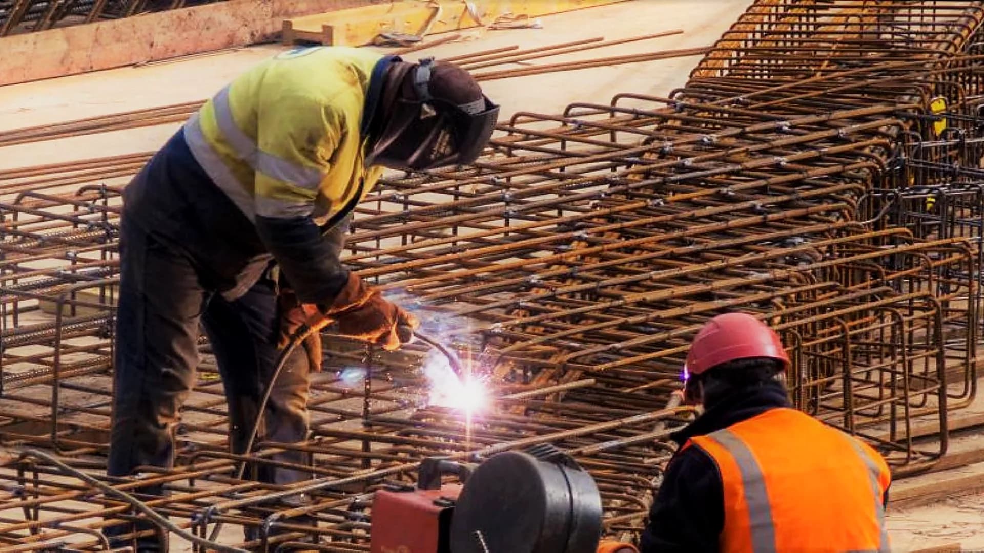 Soudeur assemblant des cages d'armatures sur un chantier béton, avec un ouvrier au premier plan